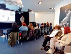 Schülergruppe lauscht einem Vortrag mit einer Bildschirmpräsentation im Hintergrund