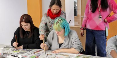eine Lehrerin zeigt zwei Schülerinnen die richtige Pinselhaltung beim Malen der asiatischen Schriftzeichen