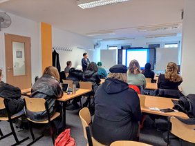 Schülergruppe von hinten, die in einem Raum sitzt und einem Vortrag mit einer Bildschirmpräsentation zuhört
