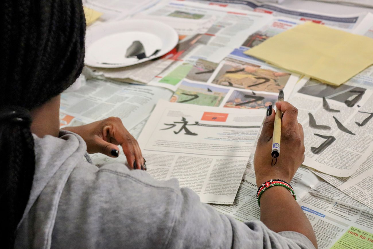Blick über die Schulter einer Schülerin, die ein ansiatisches Schriftzeichen auf eine Zeitung malt